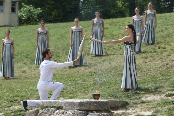 扮演最高女祭司的演員瑪麗·米娜（前右）點(diǎn)燃第一棒火炬手——希臘運(yùn)動員、東京奧運(yùn)會男子賽艇項(xiàng)目金牌得主斯特凡諾斯·恩圖斯科斯手中的火炬。新華社記者 趙丁喆 攝