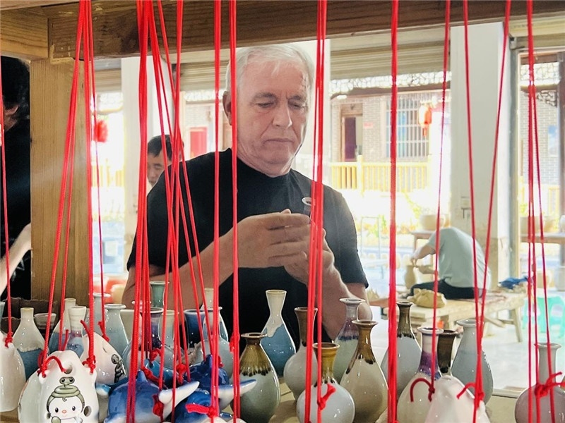 鄉(xiāng)愁如同一根根紅線，將游子與故鄉(xiāng)永遠(yuǎn)地牽連到一起?；逄账囄幕瘎?chuàng)意一條街的母氏陶藝館內(nèi)，紅線吊著陶瓶，思緒游走萬(wàn)里。摩爾多瓦“信息標(biāo)簽”通訊社社長(zhǎng)亞歷山大·塔納斯端詳著館內(nèi)展示的陶藝作品，并挑選出心儀的小擺件帶回摩爾多瓦，也帶走花茂村的“鄉(xiāng)愁”。（張亞欣攝）